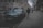 A luxury car, possibly a Rolls-Royce, is parked on a street lined with elegant, multi-story buildings. There is a black taxi cab nearby, and a person wearing a hooded jacket and beanie is walking on the sidewalk. The atmosphere is gray and somewhat moody, suggesting an overcast day in an urban setting.