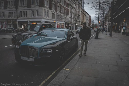 A discreet, sharply dressed personal security professional standing confidently beside a sleek black Rolls-Royce in a dimly lit London street.