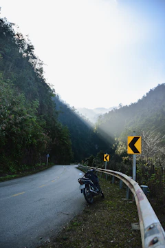 A winding mountain road with a motorcycle parked beside a viewpoint overlooking lush green valleys.