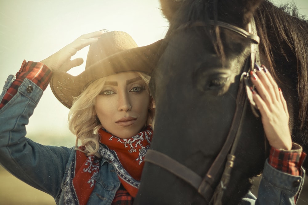 woman in blue denim jacket wearing brown cowboy hat beside black horse during daytime