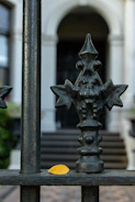 An ornate iron fence with a detailed design, featuring geometric and floral elements, stands in front of a blurred background of a building's entrance with steps. A single yellow leaf sits on the horizontal bar of the fence, providing a contrast to the dark metal.