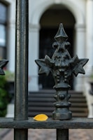 An ornate iron fence with a detailed design, featuring geometric and floral elements, stands in front of a blurred background of a building's entrance with steps. A single yellow leaf sits on the horizontal bar of the fence, providing a contrast to the dark metal.