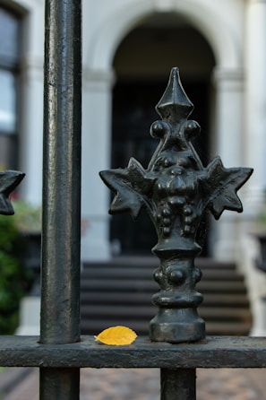 An ornate iron fence with a detailed design, featuring geometric and floral elements, stands in front of a blurred background of a building's entrance with steps. A single yellow leaf sits on the horizontal bar of the fence, providing a contrast to the dark metal.