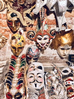 An assortment of intricately designed masks displayed against a brick wall backdrop. These include colorful Venetian-style masks with elaborate decorations, feathers, and lace. A prominent jester mask with a smiling expression is surrounded by other masks featuring gold accents and vibrant patterns.