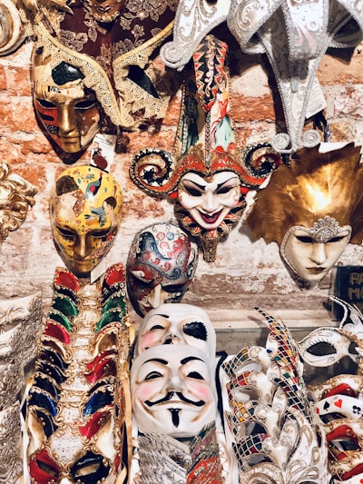 An assortment of intricately designed masks displayed against a brick wall backdrop. These include colorful Venetian-style masks with elaborate decorations, feathers, and lace. A prominent jester mask with a smiling expression is surrounded by other masks featuring gold accents and vibrant patterns.