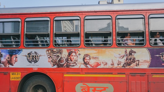 A red public bus with an advertisement on its side depicting various historical or mythical figures in elaborate costumes. The bus windows show passengers seated inside.