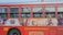 A red public bus with an advertisement on its side depicting various historical or mythical figures in elaborate costumes. The bus windows show passengers seated inside.