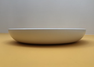 A minimalist composition featuring a white, shallow, round dish placed against a neutral, softly lit background. The dish is centered and viewed from a low angle, emphasizing its gentle curve and clean lines.