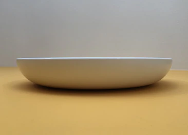 A minimalist ceramic plate with gentle curves, photographed against a pale, textured surface.