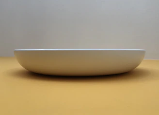 A minimalist ceramic plate with gentle curves, photographed against a pale, textured surface.