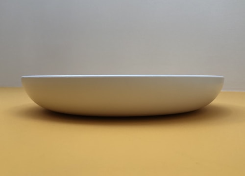 A minimalist composition featuring a white, shallow, round dish placed against a neutral, softly lit background. The dish is centered and viewed from a low angle, emphasizing its gentle curve and clean lines.