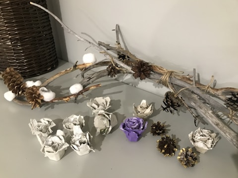 A decorative arrangement featuring a bundle of dry, rustic twigs and branches adorned with pinecones and cotton balls, positioned against a woven basket. In the foreground, creatively crafted flowers made from egg cartons are displayed, with one distinctively painted purple. Small, gold-tipped pinecones complement the scene.