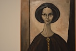 A portrait painting of a woman with distinct facial features, elongated neck, and dark hair. The background and her clothing have muted earth tones, creating a vintage and somewhat timeless appearance.