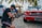 A person kneeling on cobblestone ground operates a professional video camera in front of a vintage red car. Palm trees and a sunny setting provide a tropical background, with a white car parked nearby.