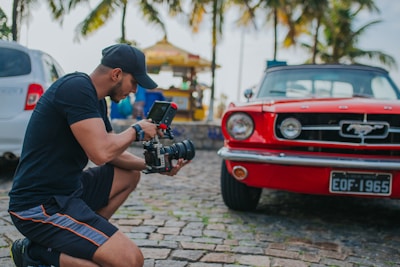 A person kneeling on cobblestone ground operates a professional video camera in front of a vintage red car. Palm trees and a sunny setting provide a tropical background, with a white car parked nearby.