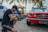 A person kneeling on cobblestone ground operates a professional video camera in front of a vintage red car. Palm trees and a sunny setting provide a tropical background, with a white car parked nearby.