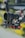 A professional camera setup with a large lens, mounted on a stabilizing rig with several red adjustment knobs and mechanical components. There is a coiled cable and various intricate details on the equipment, set against a blurred outdoor background.