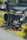 A professional camera setup with a large lens, mounted on a stabilizing rig with several red adjustment knobs and mechanical components. There is a coiled cable and various intricate details on the equipment, set against a blurred outdoor background.