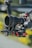 A professional camera setup with a large lens, mounted on a stabilizing rig with several red adjustment knobs and mechanical components. There is a coiled cable and various intricate details on the equipment, set against a blurred outdoor background.