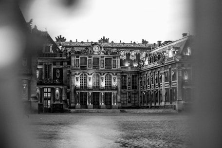 An elegant and intricate historic palace with detailed architectural features is framed by two blurred columns. The building has large, ornate windows and an elaborately decorated roofline, lit up under the soft lighting, creating a striking contrast against the cobblestone courtyard.