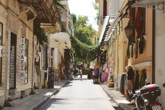 A narrow pedestrian street lined with small shops and outdoor displays, including clothing and souvenirs. Overhead, green vines create a canopy between the buildings, adding a touch of nature to the urban setting. People are casually walking and browsing the items displayed along the street.
