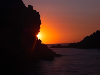 A vibrant sunset casting warm orange and pink hues over the calm ocean and rocky coastline of Noronha.