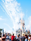 A large, magical castle with blue spires is seen in the background, surrounded by clouds of smoke and fireworks. A crowd of people, some wearing hats and Mickey Mouse ears, are gathered in the foreground, capturing the festive and lively atmosphere.