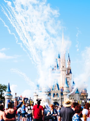 A large, magical castle with blue spires is seen in the background, surrounded by clouds of smoke and fireworks. A crowd of people, some wearing hats and Mickey Mouse ears, are gathered in the foreground, capturing the festive and lively atmosphere.