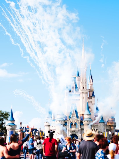 A large, magical castle with blue spires is seen in the background, surrounded by clouds of smoke and fireworks. A crowd of people, some wearing hats and Mickey Mouse ears, are gathered in the foreground, capturing the festive and lively atmosphere.