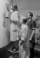 Three individuals are engaged in painting or designing on a large canvas or wall. The person closest is standing on a stool, holding a tin and a brush, and appears to be concentrating on their work. The other two are observing closely, wearing aprons or work coats, indicative of a collaborative art or creative project.