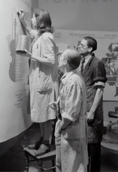 Three individuals are engaged in painting or designing on a large canvas or wall. The person closest is standing on a stool, holding a tin and a brush, and appears to be concentrating on their work. The other two are observing closely, wearing aprons or work coats, indicative of a collaborative art or creative project.