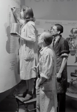 Three individuals are engaged in painting or designing on a large canvas or wall. The person closest is standing on a stool, holding a tin and a brush, and appears to be concentrating on their work. The other two are observing closely, wearing aprons or work coats, indicative of a collaborative art or creative project.
