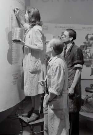 Three individuals are engaged in painting or designing on a large canvas or wall. The person closest is standing on a stool, holding a tin and a brush, and appears to be concentrating on their work. The other two are observing closely, wearing aprons or work coats, indicative of a collaborative art or creative project.