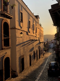 A sunlit cobblestone street flanked by a long, beige building with arched doorways and windows. The soft glow of the sunset casts long shadows, creating a warm and inviting atmosphere. Floral decorations can be seen on balconies, adding a touch of nature to the historic architecture. At the street's end, a small car is parked, enhancing the scene's vintage feel.