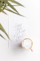 An elegant flat lay of a script-font signature card beside a cup of coffee on a clean white table.