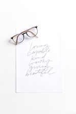 A pair of eyeglasses placed on top of a sheet of paper with handwritten words. The words include 'loving', 'capable', 'kind', 'worthy', 'giving', and 'beautiful' in a cursive style.