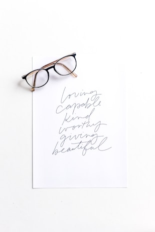 A pair of eyeglasses placed on top of a sheet of paper with handwritten words. The words include 'loving', 'capable', 'kind', 'worthy', 'giving', and 'beautiful' in a cursive style.