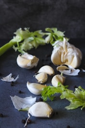 An arrangement of garlic cloves and a bunch of fresh celery is set against a dark background. The garlic is partially peeled, with pieces of the papery skin scattered around. Black peppercorns are also sprinkled across the surface.