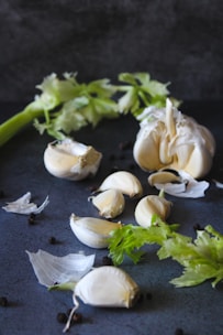 An arrangement of garlic cloves and a bunch of fresh celery is set against a dark background. The garlic is partially peeled, with pieces of the papery skin scattered around. Black peppercorns are also sprinkled across the surface.