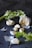 An arrangement of garlic cloves and a bunch of fresh celery is set against a dark background. The garlic is partially peeled, with pieces of the papery skin scattered around. Black peppercorns are also sprinkled across the surface.