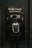 A person stands in front of an ATM machine with the sign 'Free Cash withdrawals' above it. The individual is wearing a black jacket, a black cap, and a backpack. The surrounding area is dark in color, emphasizing the bright signage and screen of the ATM.