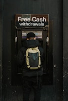 A person stands in front of an ATM machine with the sign 'Free Cash withdrawals' above it. The individual is wearing a black jacket, a black cap, and a backpack. The surrounding area is dark in color, emphasizing the bright signage and screen of the ATM.
