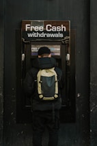 A person stands in front of an ATM machine with the sign 'Free Cash withdrawals' above it. The individual is wearing a black jacket, a black cap, and a backpack. The surrounding area is dark in color, emphasizing the bright signage and screen of the ATM.
