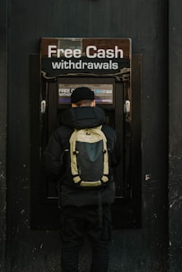 A person stands in front of an ATM machine with the sign 'Free Cash withdrawals' above it. The individual is wearing a black jacket, a black cap, and a backpack. The surrounding area is dark in color, emphasizing the bright signage and screen of the ATM.