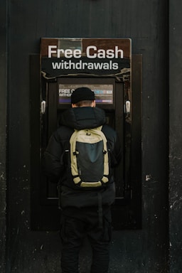 A person stands in front of an ATM machine with the sign 'Free Cash withdrawals' above it. The individual is wearing a black jacket, a black cap, and a backpack. The surrounding area is dark in color, emphasizing the bright signage and screen of the ATM.