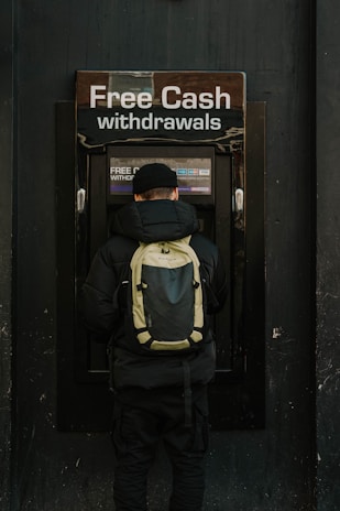 A person stands in front of an ATM machine with the sign 'Free Cash withdrawals' above it. The individual is wearing a black jacket, a black cap, and a backpack. The surrounding area is dark in color, emphasizing the bright signage and screen of the ATM.