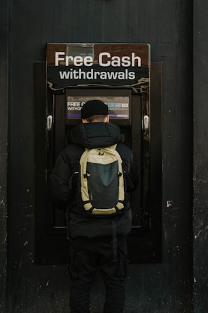A person stands in front of an ATM machine with the sign 'Free Cash withdrawals' above it. The individual is wearing a black jacket, a black cap, and a backpack. The surrounding area is dark in color, emphasizing the bright signage and screen of the ATM.