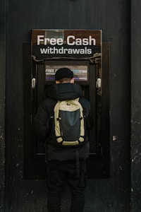 A person stands in front of an ATM machine with the sign 'Free Cash withdrawals' above it. The individual is wearing a black jacket, a black cap, and a backpack. The surrounding area is dark in color, emphasizing the bright signage and screen of the ATM.