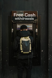 A person stands in front of an ATM machine with the sign 'Free Cash withdrawals' above it. The individual is wearing a black jacket, a black cap, and a backpack. The surrounding area is dark in color, emphasizing the bright signage and screen of the ATM.