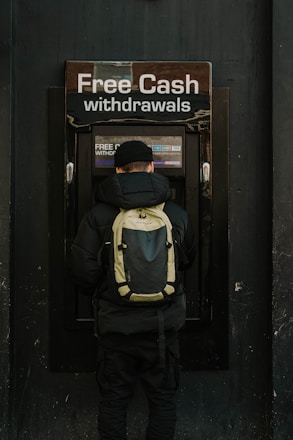A person stands in front of an ATM machine with the sign 'Free Cash withdrawals' above it. The individual is wearing a black jacket, a black cap, and a backpack. The surrounding area is dark in color, emphasizing the bright signage and screen of the ATM.